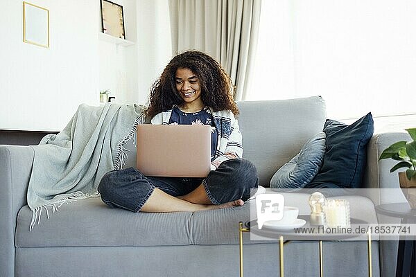 Aufgeregte  fröhliche junge schwarze Frau  die zu Hause auf dem Sofa einen Laptop benutzt  gute Nachrichten erhält  Freude empfindet  Hausaufgaben macht oder online arbeitet  lacht  sich über einen Preis freut