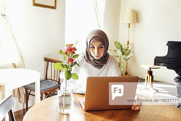 Hübsche junge Frau aus dem Nahen Osten  die einen Hijab trägt und zu Hause einen Laptop benutzt. Muslimisches Mädchen bereitet sich auf Aufnahmeprüfungen vor. Heimunterricht und Online Studium Konzept