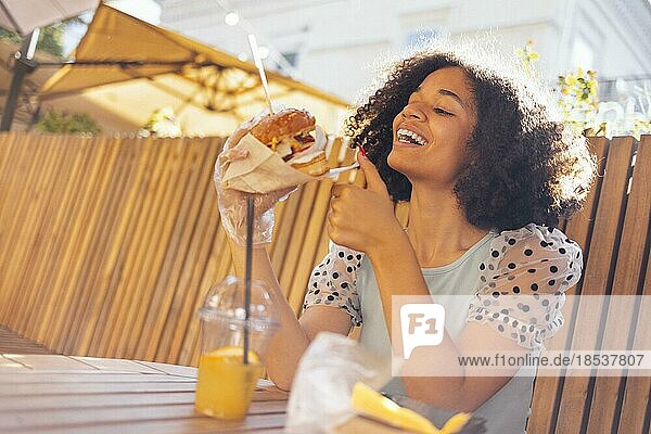Junges afrikanisches amerikanisches Mädchen ißt klassischen Burger und trinkt Limonade in der Stadt. Outdoor Frau trägt Sommerbluse mit Tupfen eatting leckeren Burger beim Sitzen auf der Terrasse. Frau in blau Verkostung Sandwich mit freudigen Blick bewundern sonnigen w