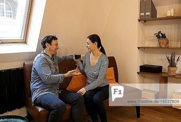 Happy husband and wife sit rest on comfortable sofa in living room enjoy tea talking  smiling. Couple relax on couch at home drink coffee  chat speak  laugh on leisure weekend