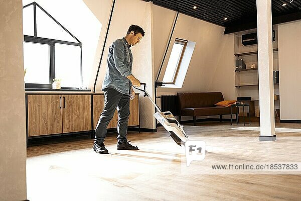 Brunette man vacuuming wooden floor in loft iving room with vacuum cleaner