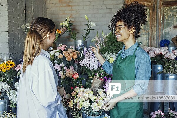 Glückliche schwarze Unternehmerin steht in einem Blumenladen und verkauft frische Blumen an Kunden. Junge blonde Mädchen kaufen einen frischen Blumenstrauß von Florist. Lächelnde afrikanische Frau Botaniker  Verkauf von Blumen