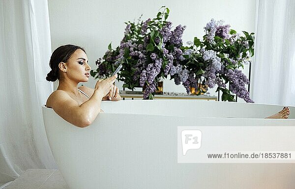 Side view portrait of a pretty asian woman typing message on mobile phone while having a bath at home