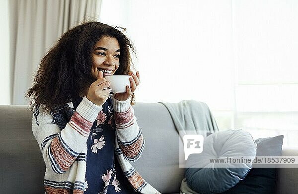 Fröhlich lächelndes afrikanisches Mädchen  das sich auf der Couch mit einer Tasse Kaffee ausruht  zu Hause ausruht  den freien Tag genießt  warme Kleidung trägt