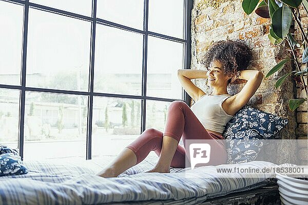Zufriedenes entspanntes afroamerikanisches Mädchen in Hauskleidung auf der Fensterbank sitzend  glückliche afroamerikanische Frau zu Hause  die sanft lächelnd zum Fenster schaut