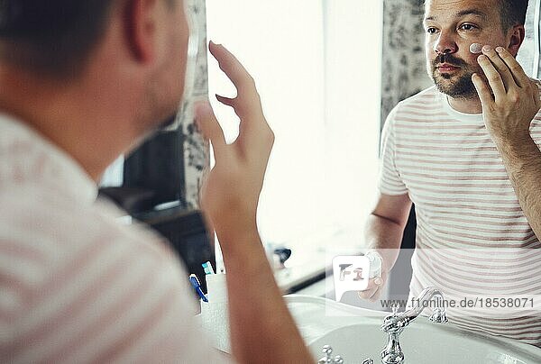 Millennial handsome man looking in mirror  applying moisturizing after shaving cream on cheeks in bathroom  head shot close up. Well groomed young guy doing skincare morning routine after showering