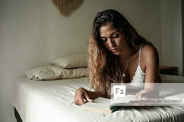 Junges Latina Mädchen mit langen Haaren liest ein Buch auf ihrem Bett in ihrem Zimmer
