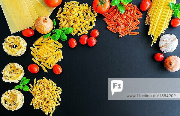 Flache Lage verschiedener ungekochter italienischer Nudeln auf dunklem Hintergrund mit Basilikumblättern  frischen Tomaten und Zwiebeln