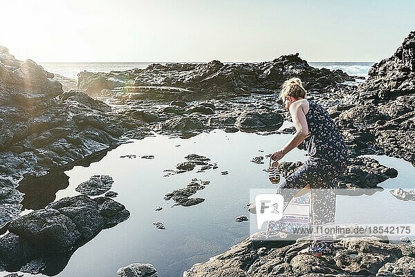 Blonde kaukasische Frau im Sommerkleid zieht ihre Schuhe am Meer an einem sonnigen Tag aus