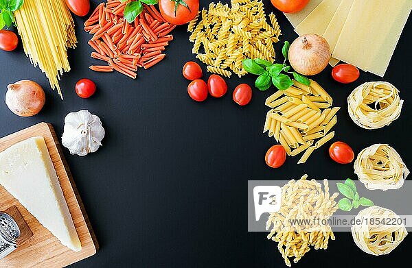 Flache Lage verschiedener ungekochter italienischer Nudeln auf dunklem Hintergrund mit Basilikumblättern  frischen Tomaten und Zwiebeln  Parmesankäse und Reibeisen