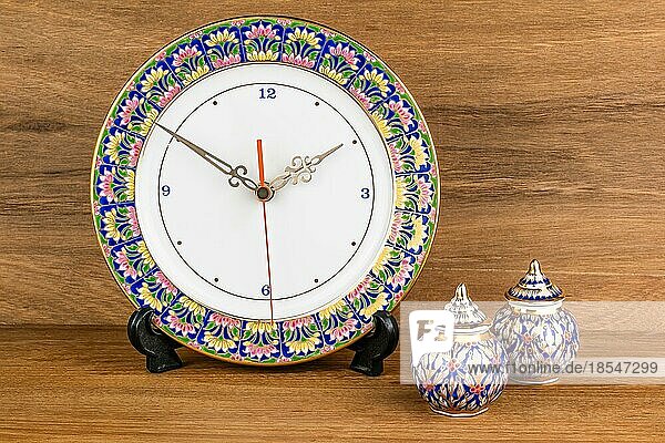 Berühmtes thailändisches Porzellan namens Benjarong Design als schöne Uhr und kleines Gefäß mit Deckel auf Holztisch. Benjarong ist Thai Porzellan Design in fünf Farbmuster
