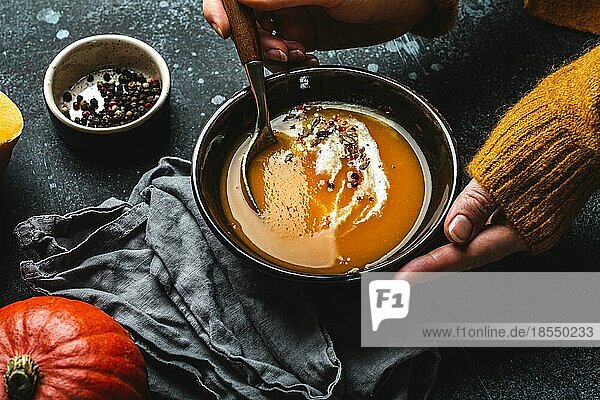 Frau in gelbem Strickpullover ißt eine Kürbiscremesuppe auf dunklem Steinhintergrund mit Löffel verziert mit geschnittenem frischem Kürbis  Ansicht von oben. Herbst gemütliches Abendessen Konzept