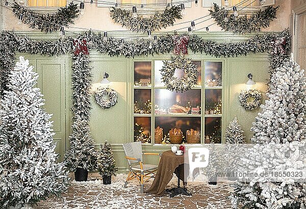 Weihnachtsatelierdekoration im Freien in pastellfarbenen Grüntönen mit einem Kaffeetisch in der Nähe der Bäckerei  Tür  Kränzen  Laternen  Girlande  Glasvitrine mit Lebkuchen  Fenster und vielen Tannenbäumen im Schnee