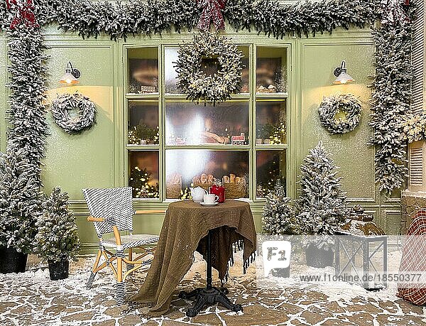 Weihnachtsatelierdekoration im Freien in pastellfarbenen Grüntönen mit einem Kaffeetisch in der Nähe der Bäckerei  Tür  Kränzen  Laternen  Girlande  Glasvitrine mit Lebkuchen  Fenster und vielen Tannenbäumen im Schnee