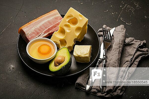 Main Keto Lebensmittel wie Butter  Olivenöl  Spiegelei  Avocado  Käse  fettes Fleisch Speck für ketogene Diät auf schwarzen Teller auf dunklen rustikalen Stein Hintergrund  Produkte für gesunde Gewichtsabnahme Draufsicht