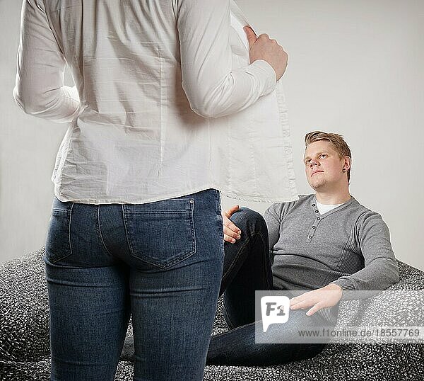 Frau zieht Bluse aus  während Mann erwartungsvoll auf Sofa sitzt