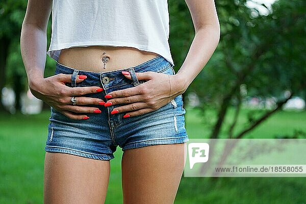 Mittelteil einer jungen Frau in Jeans-Hotpants oder Booty-Shorts  Bauchnabelpiercing und orangefarbenem Nagellack. Modetrend