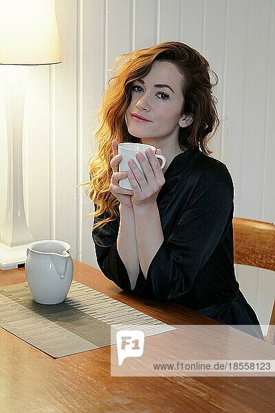 Junge Frau im Morgenmantel trinkt eine Tasse Kaffee am Küchentisch und genießt die Ruhe zu Hause
