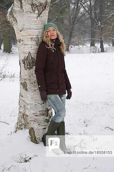 Blonde Frau lehnt an einer Birke in einer verschneiten Winterlandschaft