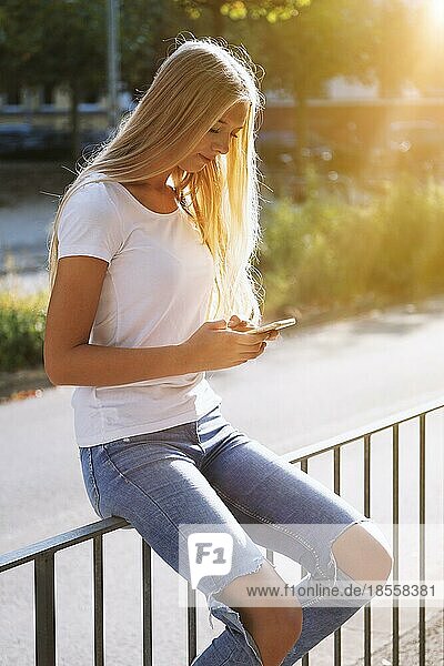 Städtischen Teenager-Mädchen mit ihrem Smartphone oder Handy  während auf dem Geländer durch die Stadt Straße sitzen - candid echte Menschen Technologie-Konzept - Gegenlicht mit Sonne Flare Lichtleck