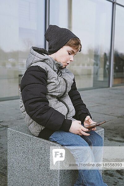 7-jähriger Junge  der mit seinem Smartphone spielt und draußen in Winterjacke und Strickmütze sitzt