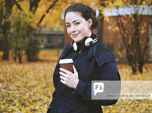Trendige junge Frau mit Kopfhörern um den Hals und Coffee-to-go-Becher in der Hand  die an einem kalten Herbsttag im Park spazieren geht