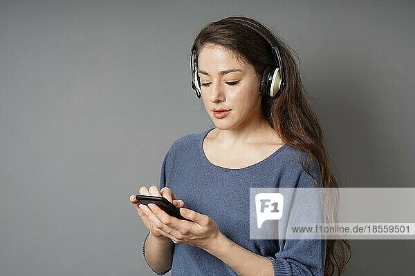 Junge Frau in den 20ern hört Musik mit drahtlosen Bluetooth-Over-Ear-Kopfhörern und mobilem Smartphone - grauer Hintergrund mit Kopierraum