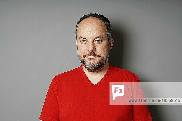 middle aged man in his 40s wearing red t-shirt with short dark hair and graying beard smirking - gray wall background with copy space