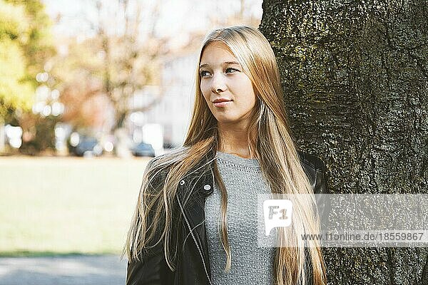Verträumtes Teenager-Mädchen mit langen blonden Haaren und Lederjacke lehnt an einem sonnigen Frühlingstag an einem Baum