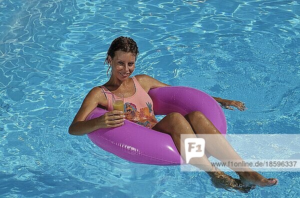 Junge Frau auf rosa Schwimmreifen mit Getränk  Glas  Cocktail in der Hand  schwimmt im swimmingpool  klares blaues Wasser