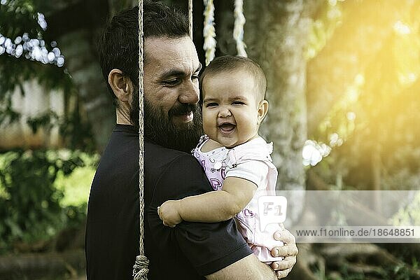 Porträt von Vater und Baby lächelnd und Blick in die Kamera sitzen in einer Hängematte. Vatertag Konzept. Sonnenuntergang Licht