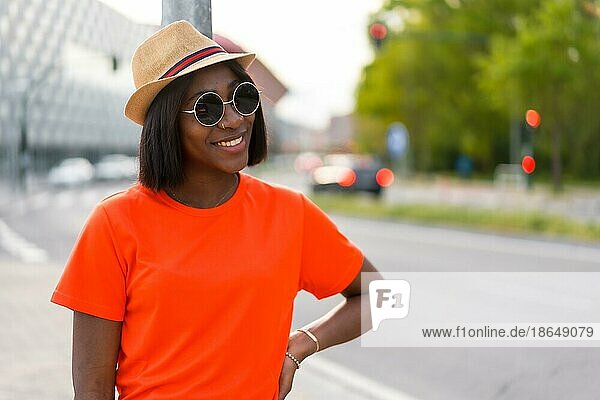 Sommerspaß: Junge schwarze Frau mit Sonnenbrille und Hut  trägt orangefarbenes TShirt lächelnd in der Stadt  Lifestylefotos