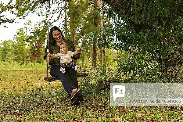 Glücklich fröhlich niedlichen Baby Mädchen sitzt auf einer Schaukel mit ihrer Mutter spielen auf dem Spielplatz