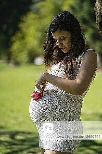 Schwangere lateinamerikanische Frau hält Babyschuhe auf ihrem Bauch in einem Park