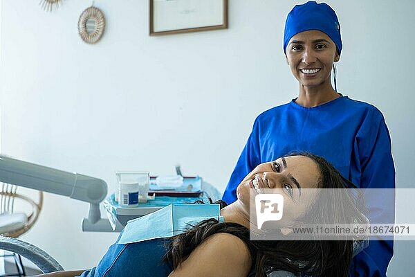 Schöne junge Frau lächelt auf dem Zahnarztstuhl. Sie und ihr Arzt schauen zufrieden mit dem Ergebnis der kieferorthopädischen Behandlung in die Kamera