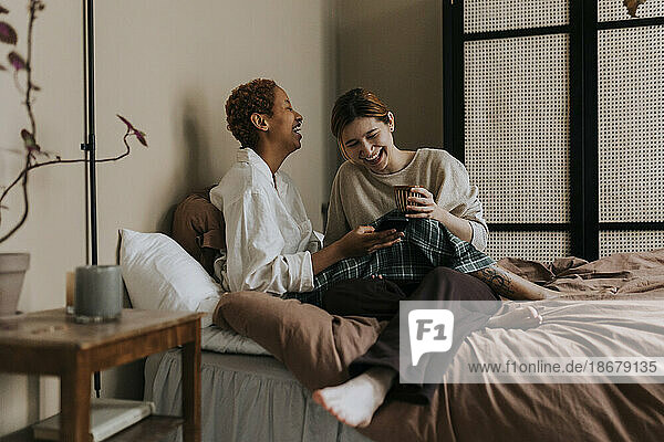 Glückliches nicht-binäres Paar teilt sich ein Smartphone  während es zu Hause auf dem Bett sitzt