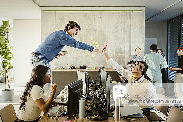 Fröhliche Unternehmer geben sich die Hand  während Kollegen in einem kreativen Büro am Schreibtisch sitzen
