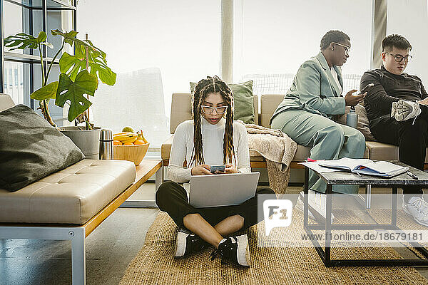 Programmierer  der ein Mobiltelefon benutzt  während er mit seinem Laptop auf dem Teppich im Kreativbüro sitzt