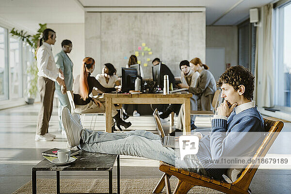 Seitenansicht eines jungen Geschäftsmannes  der auf einem Stuhl einen Laptop benutzt  während im Hintergrund Kollegen im Kreativbüro zu sehen sind