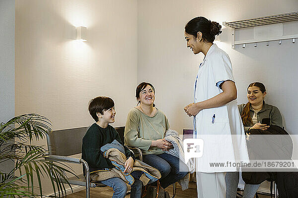 Seitenansicht einer Mitarbeiterin im Gesundheitswesen  die einen Laborkittel trägt und mit einem Jungen spricht  der im Wartezimmer eines Krankenhauses auf einem Stuhl sitzt