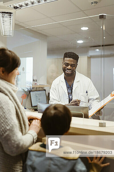 Lächelnder männlicher Rezeptionist im Gespräch mit Mutter und Sohn durch einen transparenten Schild in einer Klinik