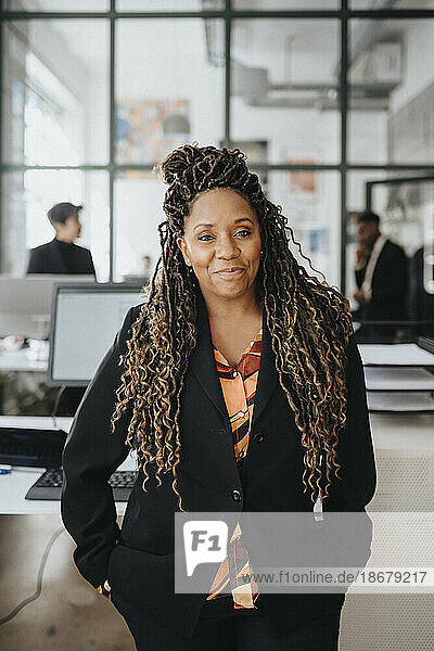 Porträt einer lächelnden reifen Geschäftsfrau mit Händen in den Taschen  die im Büro steht