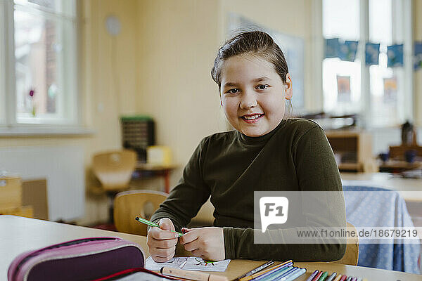 Portrait of smiling female pupil sitting at desk in classroom