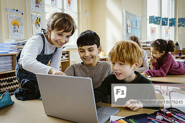 Happy schoolboys using laptop together at desk in classroom