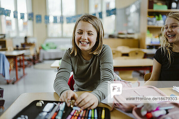 Happy schoolgirl laughing while sitting with friend at desk in classroom