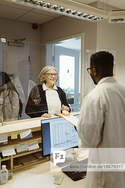 Lächelnde ältere Frau im Gespräch mit einem männlichen Rezeptionisten durch einen transparenten Schild in einer Klinik
