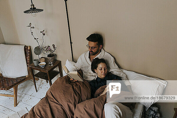 Hoher Blickwinkel auf einen Mann  der ein Buch liest  während eine nicht-binäre Person ein Mobiltelefon auf dem Bett zu Hause benutzt