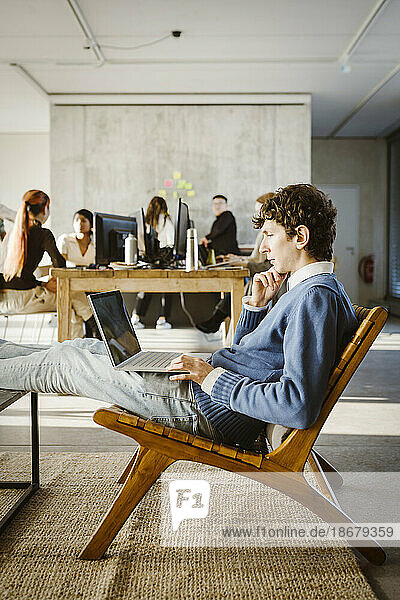 Junger Geschäftsmann  der einen Laptop auf einem Stuhl benutzt  während seine Kollegen im Hintergrund im Kreativbüro sitzen