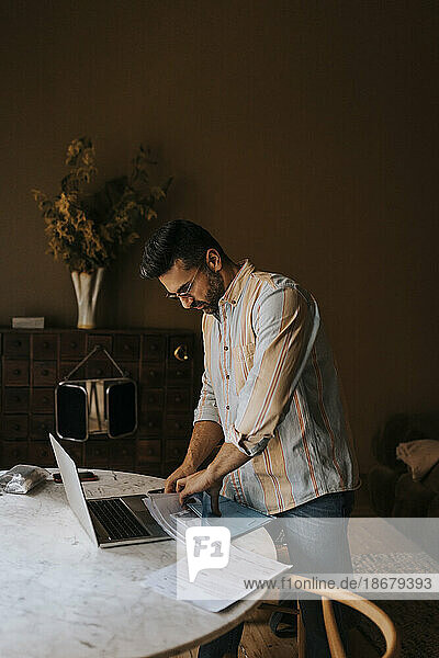Mann sortiert Dokumente in der Nähe des Laptops auf dem Tisch  während er zu Hause steht