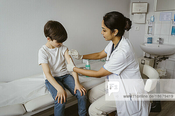 Female pediatrician disinfecting boy's arm sitting on bed at healthcare center
